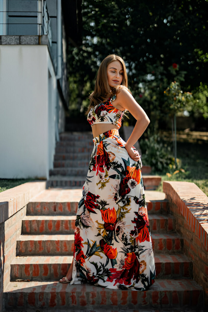 A stylish woman in a matching floral crop top and maxi skirt is posing outdoors on brick stairs in sunlight.