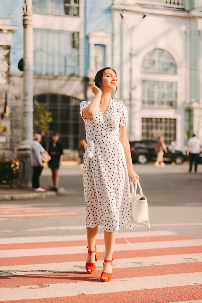 Woman wearing a vintage-style white polka dot dress with red heels and white handbag, ideal for a tea party.